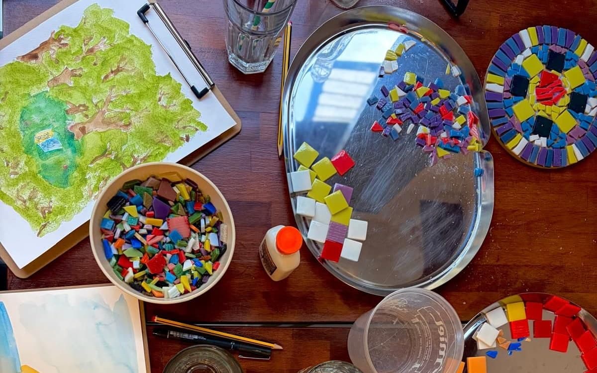 A table mid-session: watercolour on a clipboard, a bowl of tesserae and mosaic coasters in progress